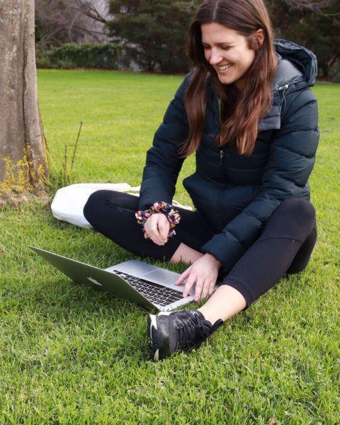 A woman wearing a black puffer jacket and leggings is sitting on grass with a laptop in front of her