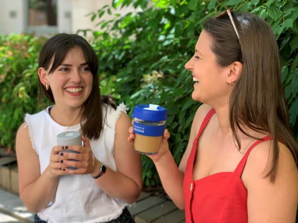 Two women are sitting down laughing at each other with two different types of reusable coffee cups in their hands.The woman on the right side is holding a clear cup with royal blue lid printed with the University of Melbourne logo on the side.