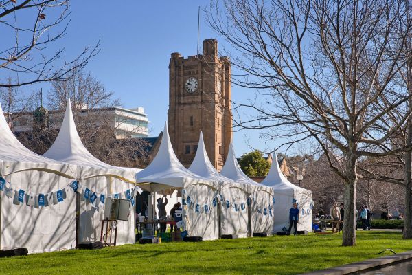 Row of white marquees with University of Melbourne-branded bunting hanging across, on a green lawn at an Open day event.
