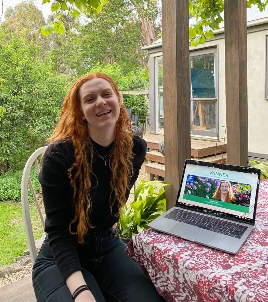 Woman with long red hair sits smiling at a table with a laptop on it. The laptop has an image of her face with the word 'winner' above.