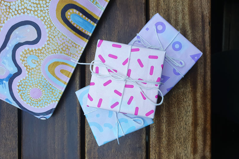 Birds eye view of three small square presents stacked on top of each other on a dark wooden table. They are each wrapped in blue, pink and purple patterned paper with white string.