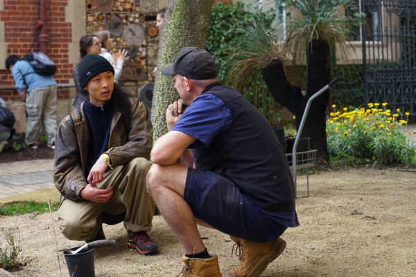 Two men crouch down talking to each other in a gravel area of a garden. 