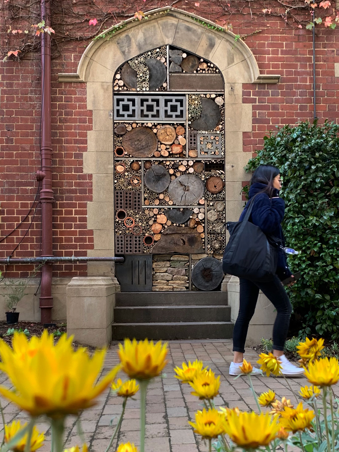 Photo of a brick doorway filled with logs, ends of bamboo, bricks and smaller branches. A woman is pictured having just walked past and there are yellow flowers in the foreground.
