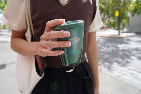 Close up of a girl holding a dark green reusable coffee cup with text reading 'Choose to Reuse @ UniMelb' and 'green caffeen' on it in white. The girl is standing on a footpath on a sunny street with trees in the background.