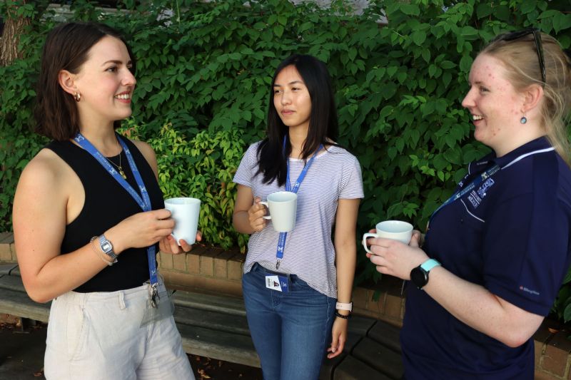 Three women wearing University of Melbourne Lanyards holding coffee mugs in in front of a bush